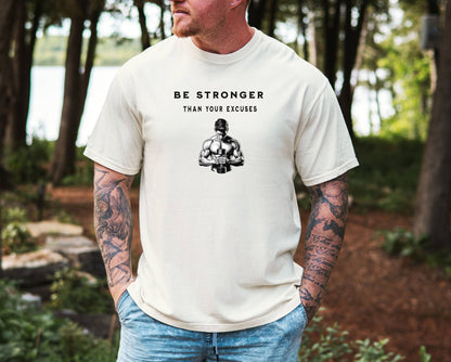 Person wearing an ivory T-shirt featuring the motivational phrase “Be Stronger Than Your Excuses” in bold black lettering, with a graphic of a muscular figure holding a dumbbell below the text. Perfect for gym-goers, fitness enthusiasts, and athletes, this empowering workout shirt inspires determination, discipline, and strength training focus.