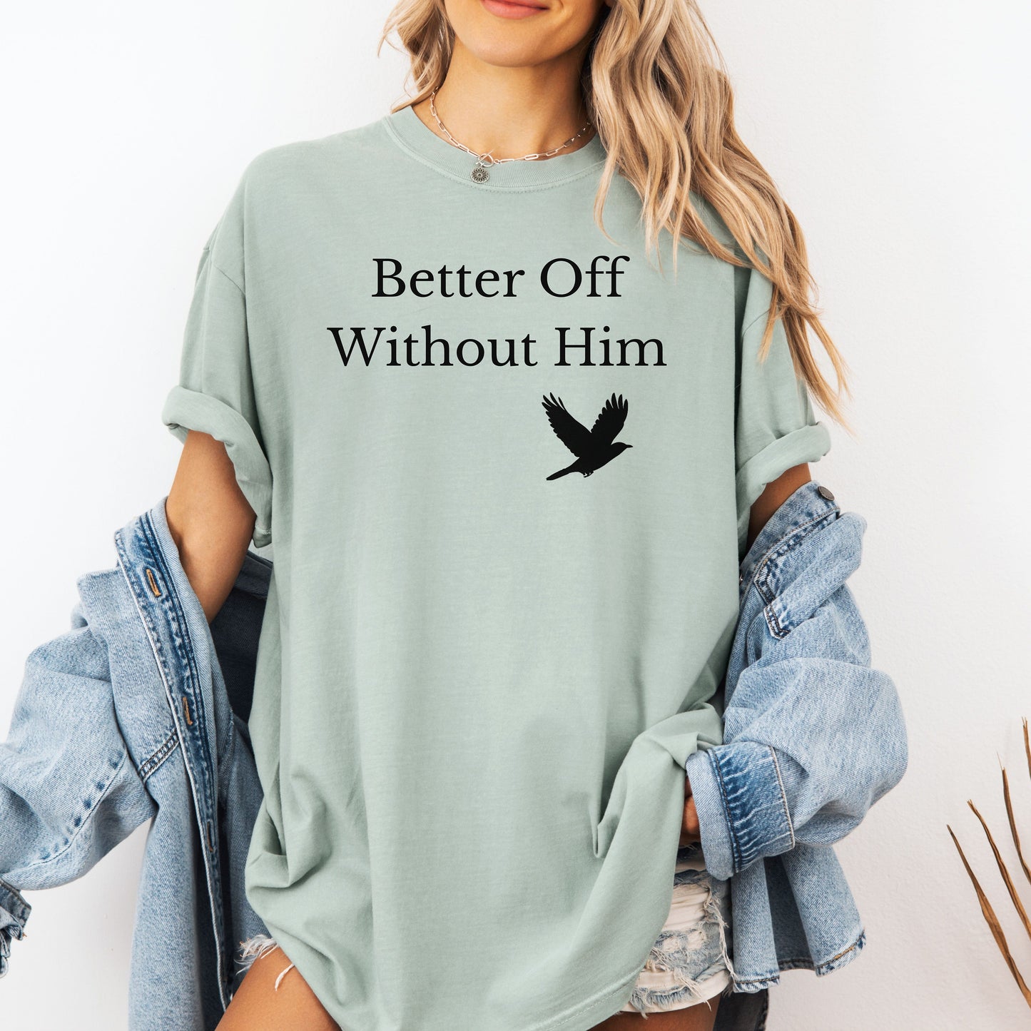 Bay light green T-shirt with the bold black phrase "Better Off Without Him" printed in a clean serif font, featuring a simple silhouette of a flying black bird that symbolizes freedom and transformation beneath the text. 