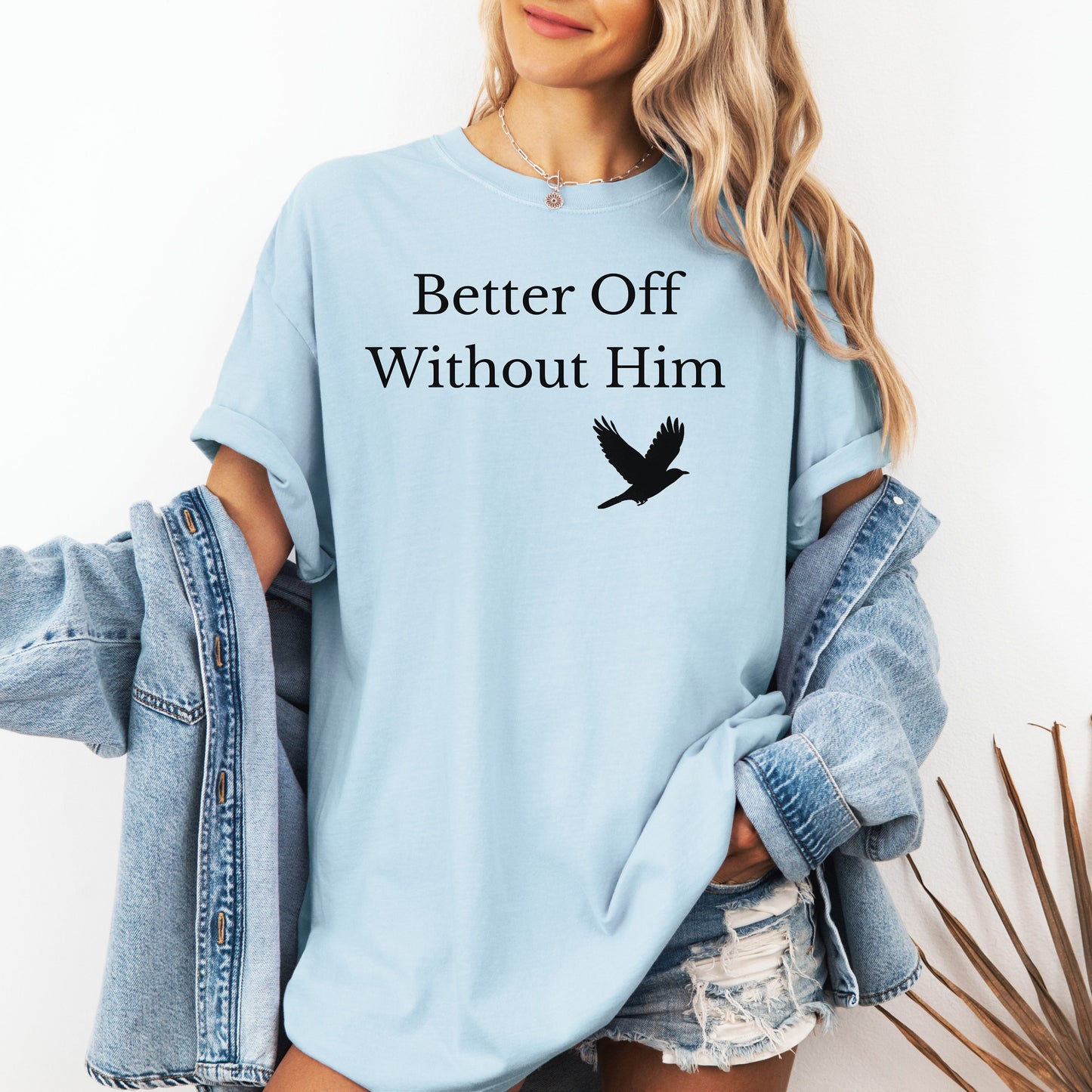 Chambray light blue T-shirt with the bold black phrase "Better Off Without Him" printed in a clean serif font, featuring a simple silhouette of a flying black bird that symbolizes freedom and transformation beneath the text. 