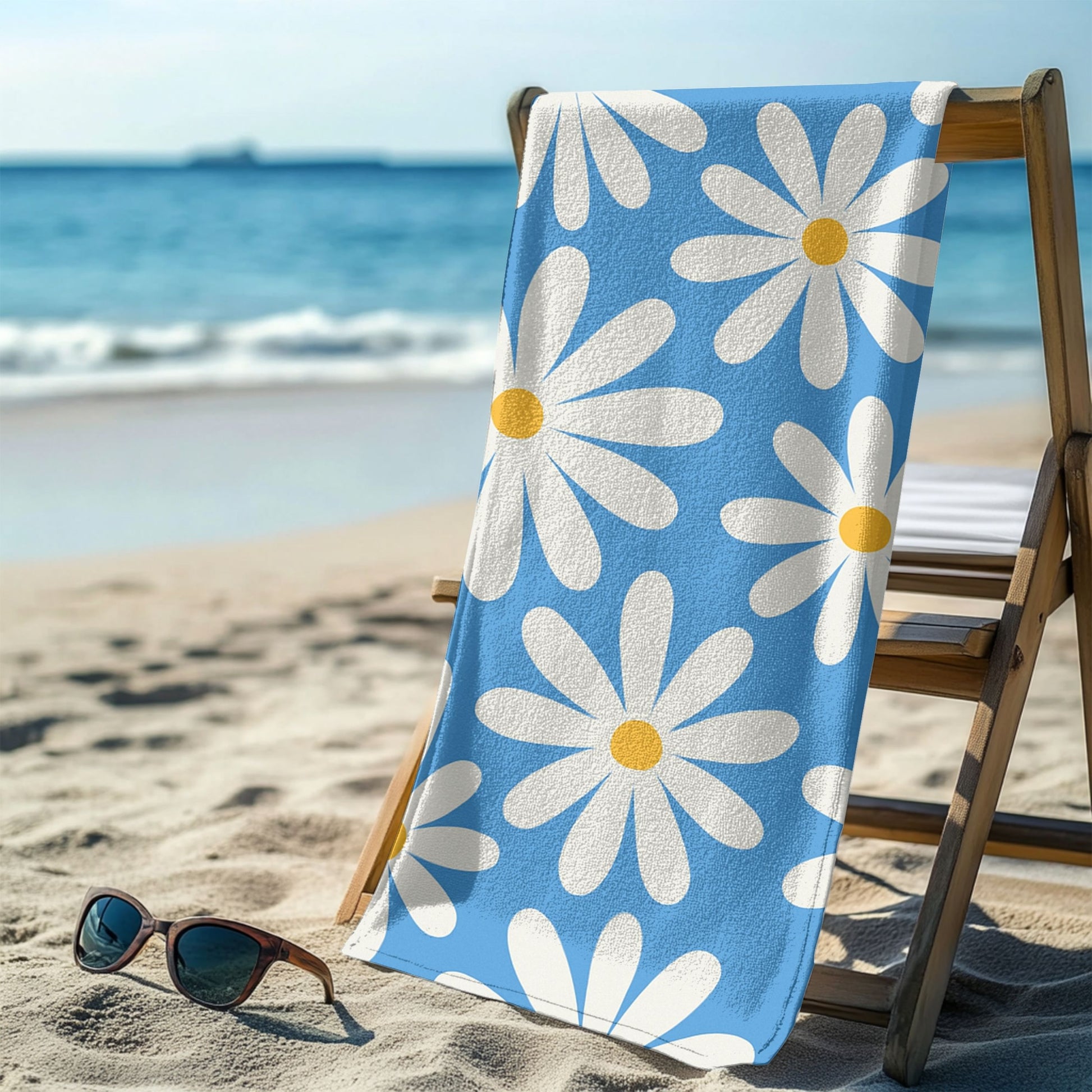 A vibrant blue beach towel featuring a retro daisy pattern with large white petals and yellow centers hanging over a beach chair. Set against a sunny beach and ocean backdrop, this playful floral design brings cheerful vintage vibes, perfect for summer days, beach trips, and poolside lounging.