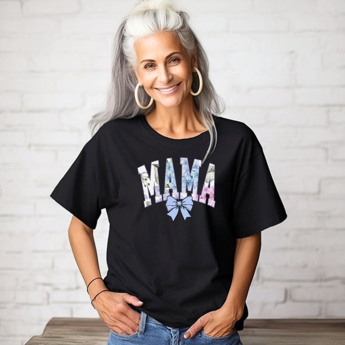 Person wearing a black T-shirt featuring the word “MAMA” in large block letters filled with a delicate blue hydrangea floral pattern, accented with soft green and pink tones. Below the text is a pastel blue coquette bow, adding a sweet, feminine touch.  This spring-inspired graphic tee blends floral elegance with playful charm, perfect for fashionable moms.