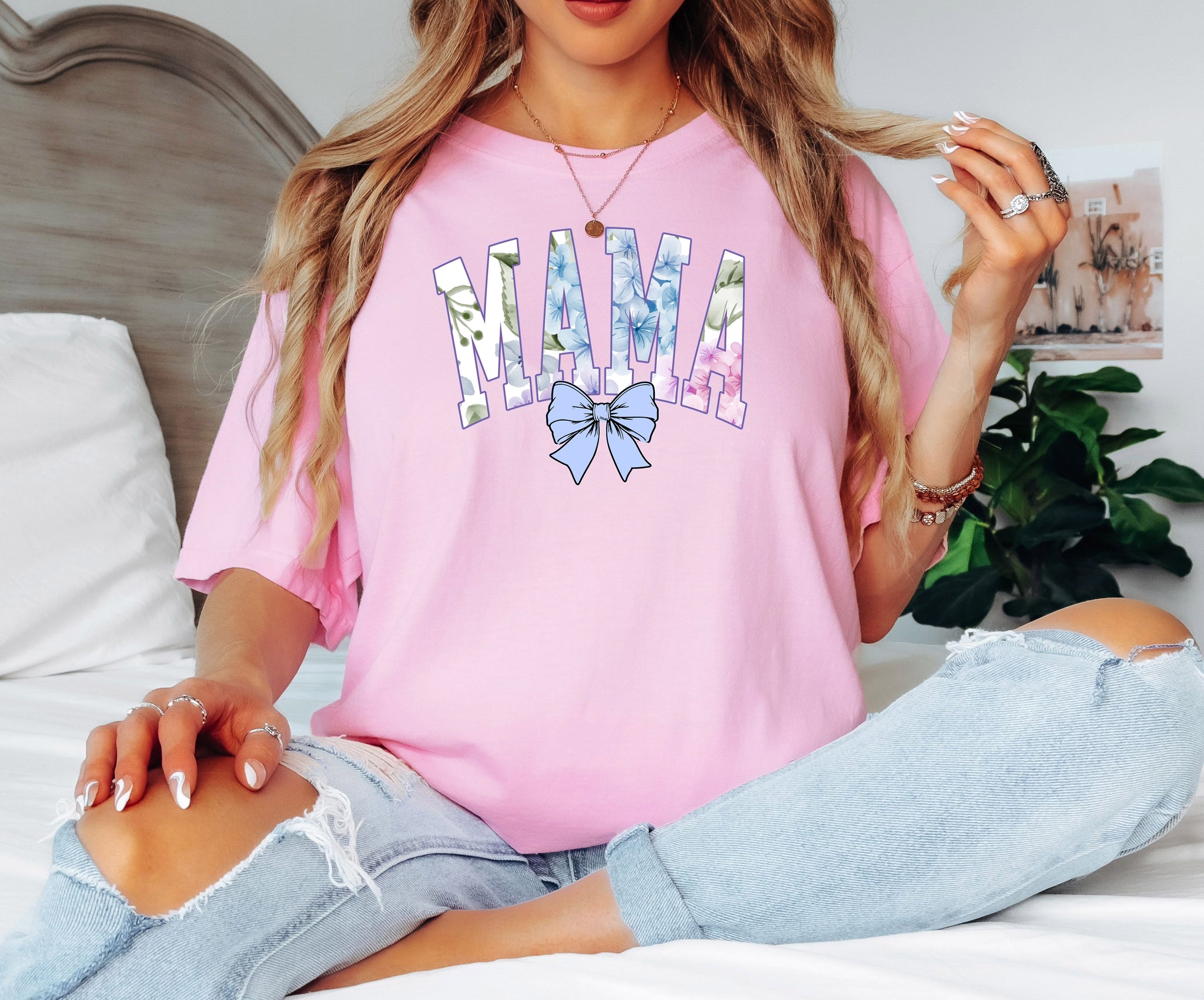 Person wearing a blossom pink T-shirt featuring the word “MAMA” in large block letters filled with a delicate blue hydrangea floral pattern, accented with soft green and pink tones. Below the text is a pastel blue coquette bow, adding a sweet, feminine touch.  This spring-inspired graphic tee blends floral elegance with playful charm, perfect for fashionable moms.