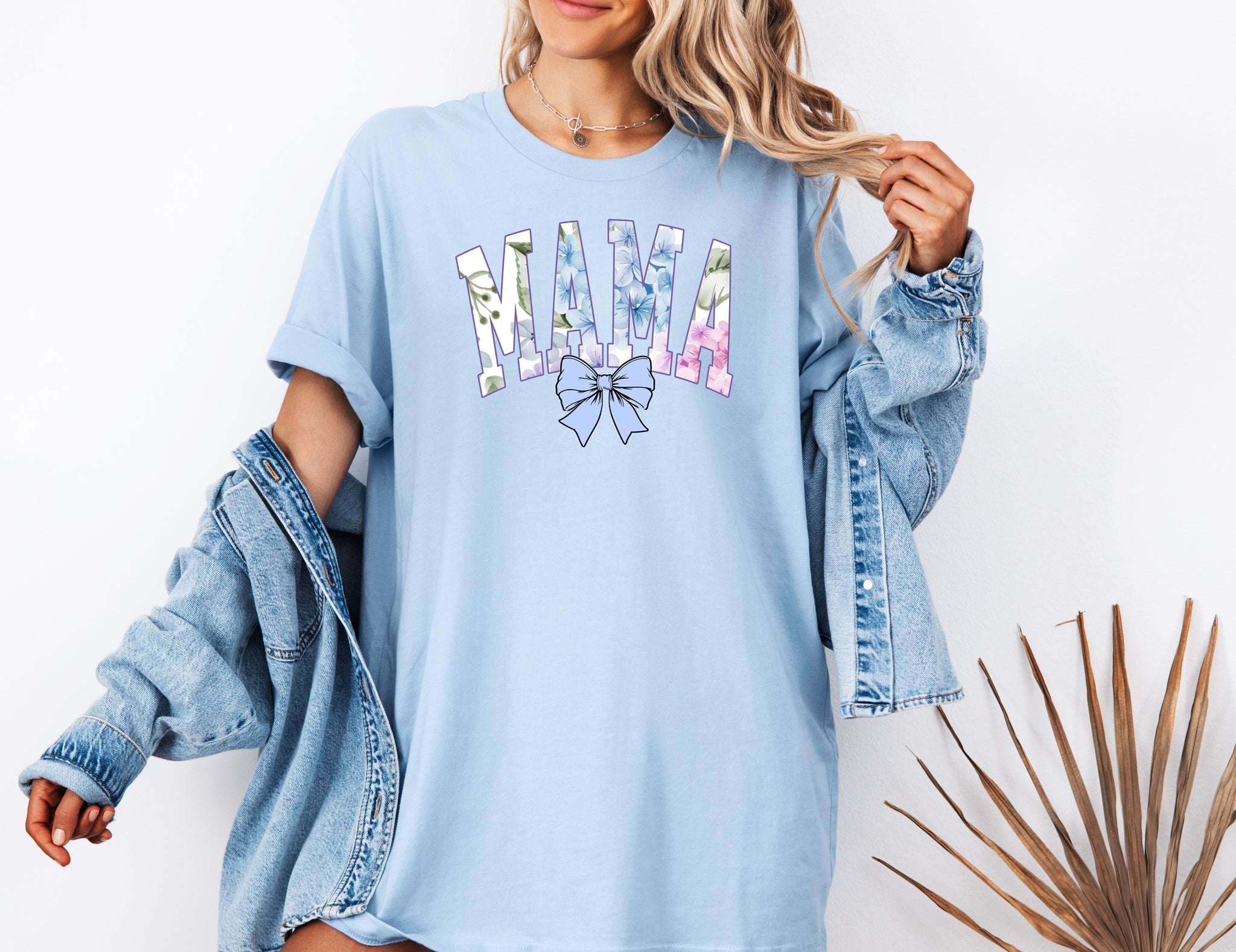 Person wearing a chambray light blue T-shirt featuring the word “MAMA” in large block letters filled with a delicate blue hydrangea floral pattern, accented with soft green and pink tones. Below the text is a pastel blue coquette bow, adding a sweet, feminine touch.  This spring-inspired graphic tee blends floral elegance with playful charm, perfect for fashionable moms.