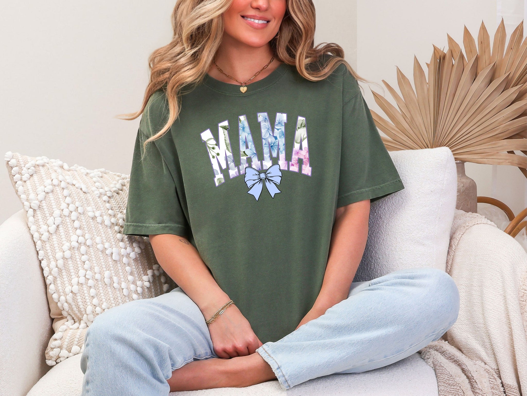 Person wearing a hemp color T-shirt featuring the word “MAMA” in large block letters filled with a delicate blue hydrangea floral pattern, accented with soft green and pink tones. Below the text is a pastel blue coquette bow, adding a sweet, feminine touch.  This spring-inspired graphic tee blends floral elegance with playful charm, perfect for fashionable moms.