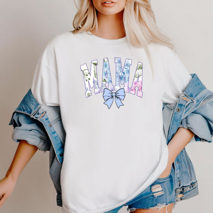 Person wearing a classic white T-shirt featuring the word “MAMA” in large block letters filled with a delicate blue hydrangea floral pattern, accented with soft green and pink tones. Below the text is a pastel blue coquette bow, adding a sweet, feminine touch.  This spring-inspired graphic tee blends floral elegance with playful charm, perfect for fashionable moms.
