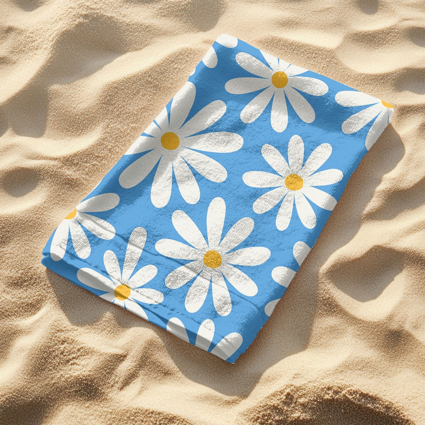 A vibrant blue beach towel featuring a retro daisy pattern with large white petals and yellow centers folded laying on the sand.  This playful floral design brings cheerful vintage vibes, perfect for summer days, beach trips, and poolside lounging.