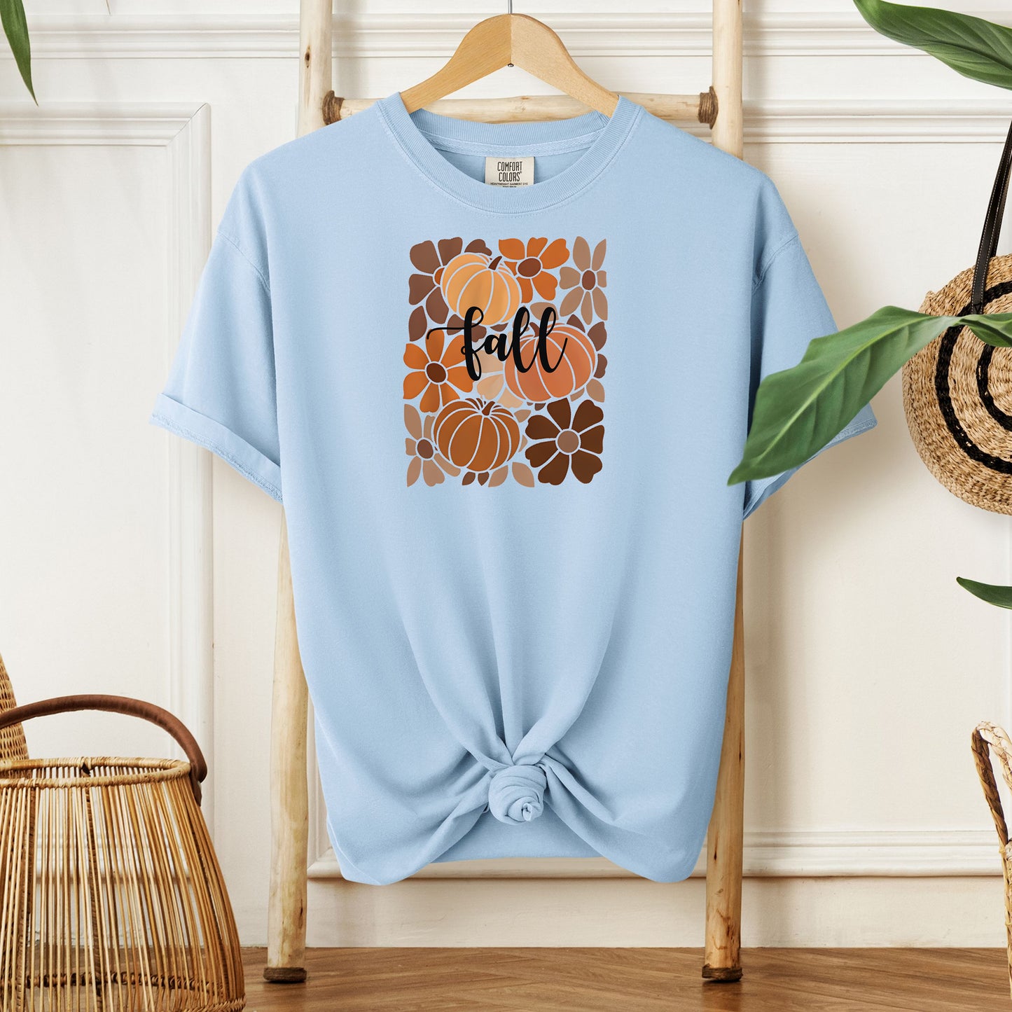 Chambray light blue T-shirt featuring a boho-inspired fall design with hand-drawn pumpkins and flowers in warm autumn tones. The word "fall" is written in elegant script across the center of the graphic, framed by a mosaic-style floral and pumpkin pattern. Perfect for seasonal style and cozy autumn vibes.