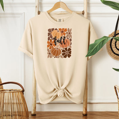 Ivory T-shirt featuring a boho-inspired fall design with hand-drawn pumpkins and flowers in warm autumn tones. The word "fall" is written in elegant script across the center of the graphic, framed by a mosaic-style floral and pumpkin pattern. Perfect for seasonal style and cozy autumn vibes.