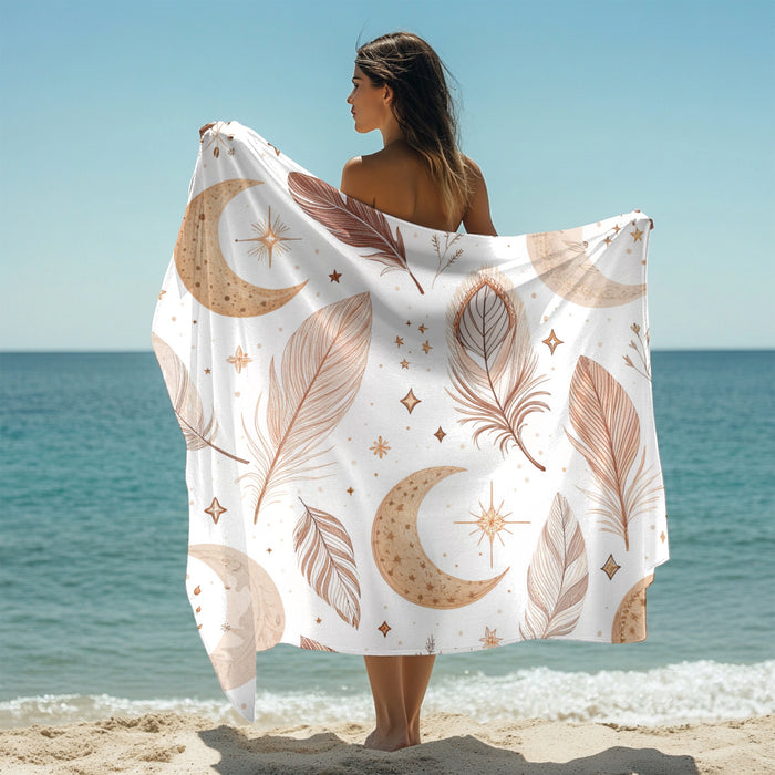 Woman holding up a white beach towel across her back featuring a boho-style design with earthy-toned feathers, crescent moons, stars, and celestial accents. Set against a sunny ocean backdrop, this mystical and artistic towel brings dreamy, free-spirited vibes—perfect for beach days, festivals, or mindful relaxation.