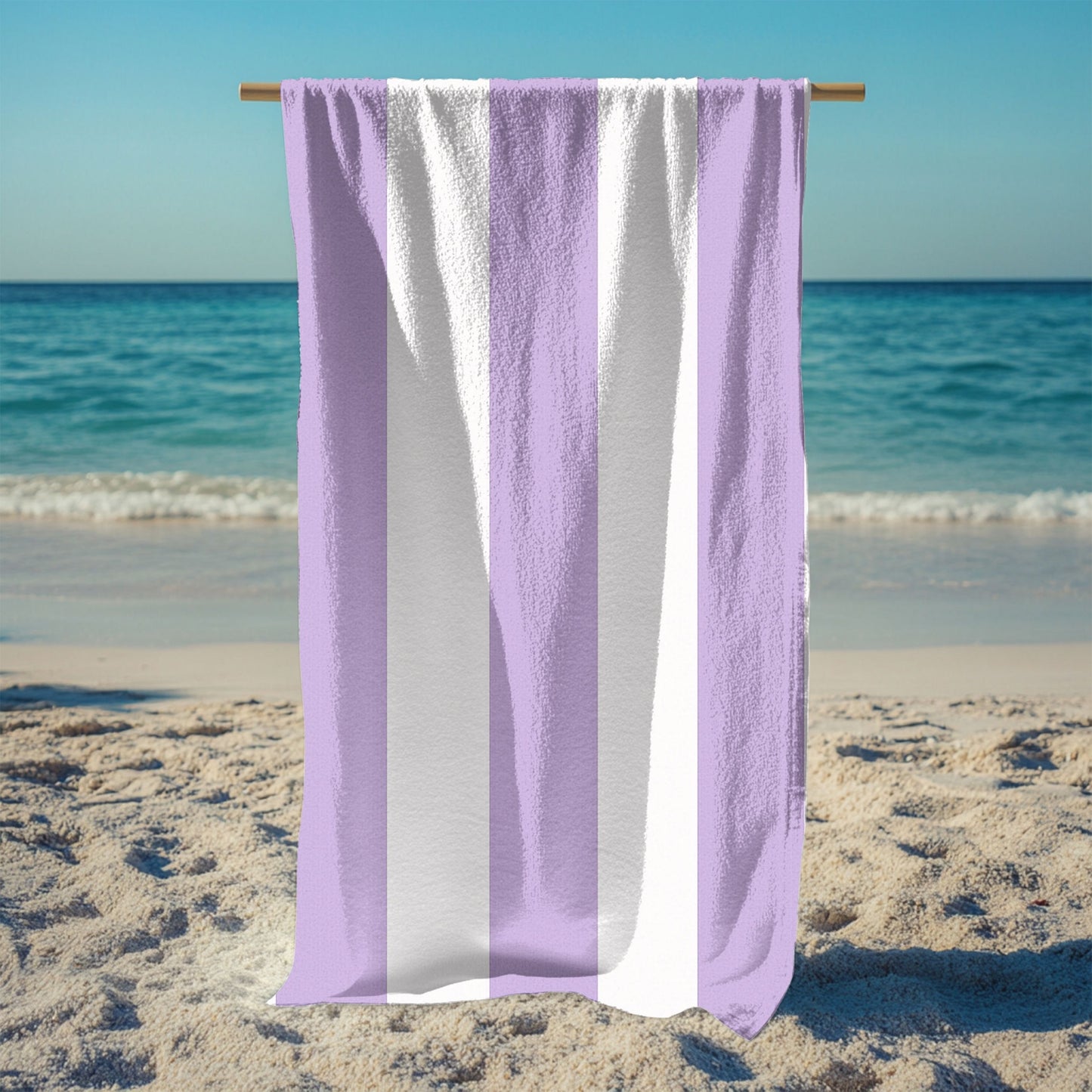 A stand holding up a beach towel with bold vertical lavender and white stripes, standing on a sunny beach with the ocean in the background. The fresh, classic striped design brings a soft and stylish touch to any seaside escape, perfect for lounging, drying off, or making a beachside statement.