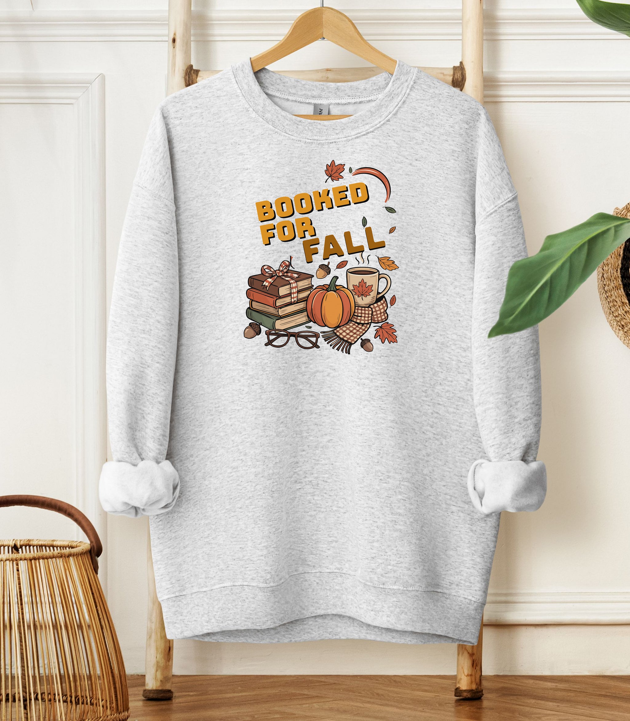 Ash Gildan sweatshirt featuring a cozy autumn-themed “Booked For Fall” design with stacked books, pumpkin, coffee mug, scarf, glasses, and falling leaves in warm fall tones. Perfect for book lovers who embrace sweater weather, pumpkin spice, and cozy reading sessions during the autumn season.