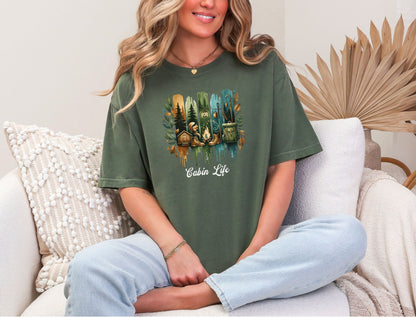 Person wearing a hemp color T-shirt with a “Cabin Life” design featuring a colorful, watercolor-style vertical stripe background in earthy tones. The artwork includes pine trees, a log cabin, a campfire with boots, a lantern, and a backpack framed by autumn leaves. A rustic, outdoorsy design perfect for nature lovers, campers, and cabin getaways.