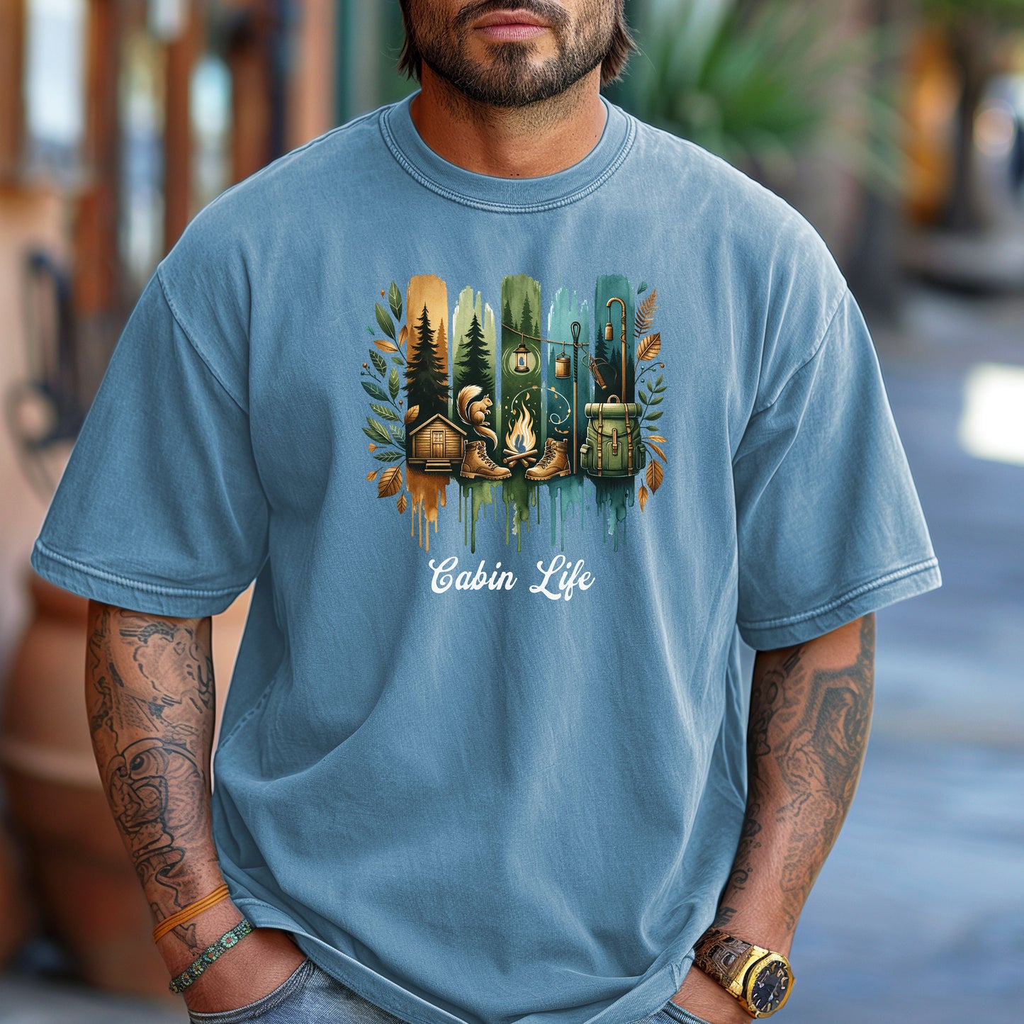 Person wearing a Washed Denim T-shirt with a “Cabin Life” design featuring a colorful, watercolor-style vertical stripe background in earthy tones. The artwork includes pine trees, a log cabin, a campfire with boots, a lantern, and a backpack framed by autumn leaves. A rustic, outdoorsy design perfect for nature lovers, campers, and cabin getaways.