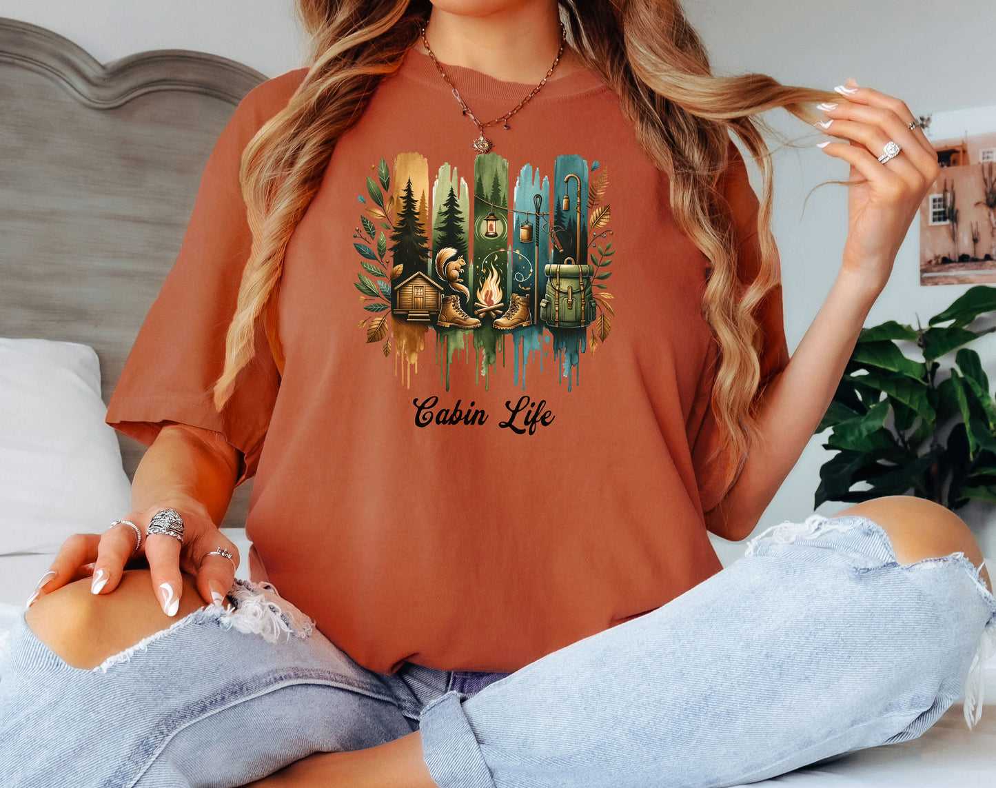 Person wearing a yami T-shirt with a “Cabin Life” design featuring a colorful, watercolor-style vertical stripe background in earthy tones. The artwork includes pine trees, a log cabin, a campfire with boots, a lantern, and a backpack framed by autumn leaves. A rustic, outdoorsy design perfect for nature lovers, campers, and cabin getaways.