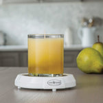 Glass jar candle sitting on a white electric candle warmer plate labeled "Candle Warmers Etc." on a kitchen countertop, with fresh pears and kitchenware blurred in the background. Ideal for safely melting candles without a flame, releasing fragrance throughout the room.