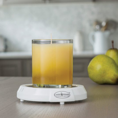 Glass jar candle sitting on a white electric candle warmer plate labeled "Candle Warmers Etc." on a kitchen countertop, with fresh pears and kitchenware blurred in the background. Ideal for safely melting candles without a flame, releasing fragrance throughout the room.