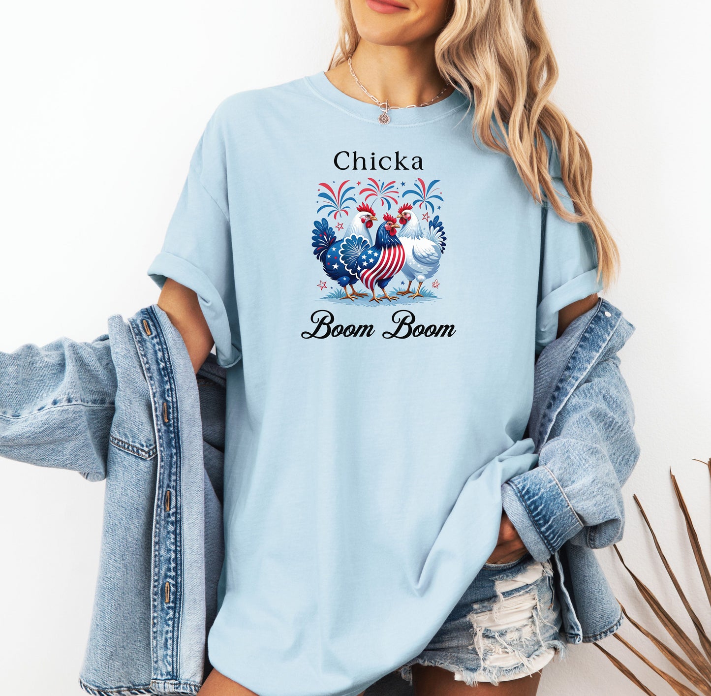 Chambray light blue t-shirt featuring a playful 'Chicka Boom Boom' design with three patriotic chickens, one decorated in red, white, and blue stars and stripes, and the other in white, surrounded by colorful fireworks. A third chicken is decorated in red and white stars on a blue body.  A fun and festive shirt perfect for 4th of July, barbecues, summer parties, and casual patriotic celebrations.