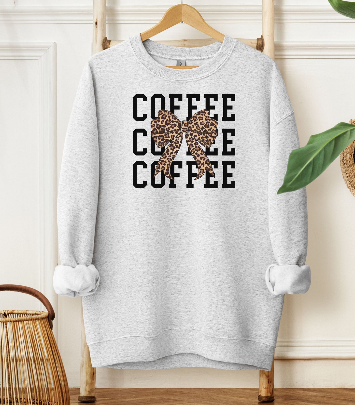 Ash Gildan sweatshirt featuring a bold black triple “COFFEE” text stacked vertically, accented with a stylish leopard-print coquette bow at the center. A trendy and playful design that blends coffee love with fashionable flair—perfect for cozy mornings and confident, cute style.