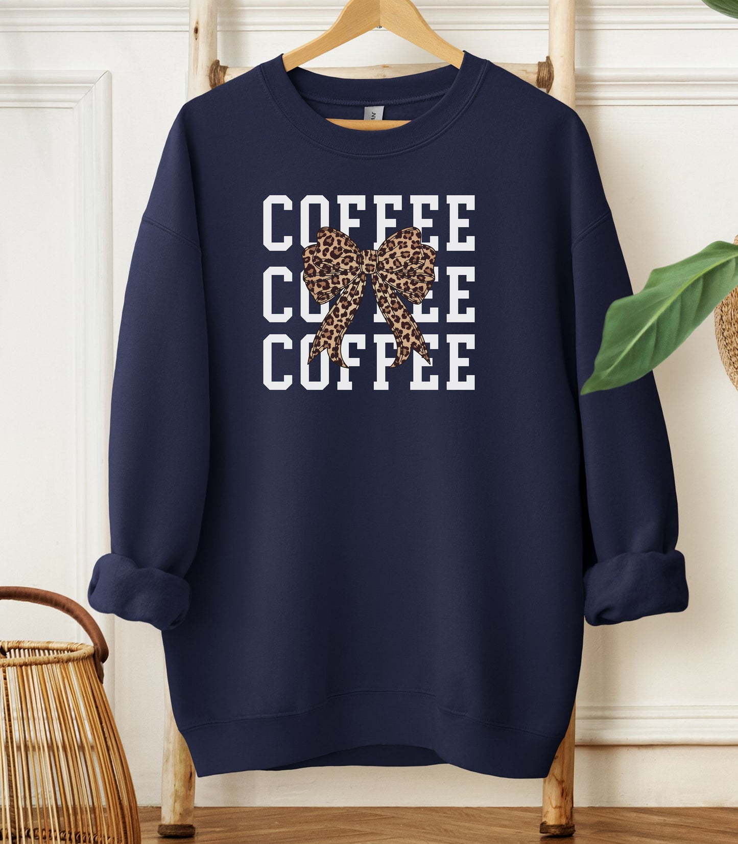 Navy Gildan sweatshirt featuring a bold white triple “COFFEE” text stacked vertically, accented with a stylish leopard-print coquette bow at the center. A trendy and playful design that blends coffee love with fashionable flair—perfect for cozy mornings and confident, cute style.