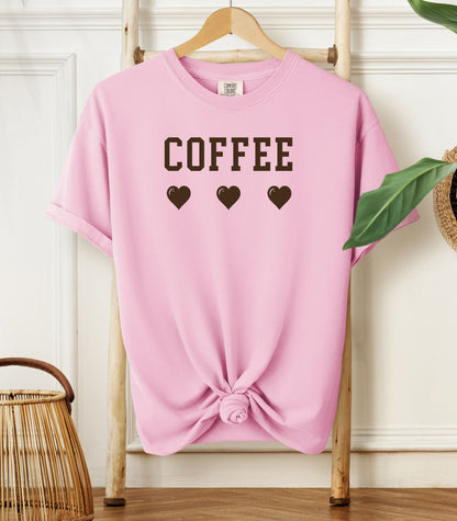 Blossom Pink Comfort Colors t-shirt featuring bold brown varsity-style “COFFEE” text above three matching heart icons. Simple and cute design that expresses love for coffee with a soft, cozy aesthetic—perfect for casual wear, coffee dates, or gifting to your favorite caffeine enthusiast.