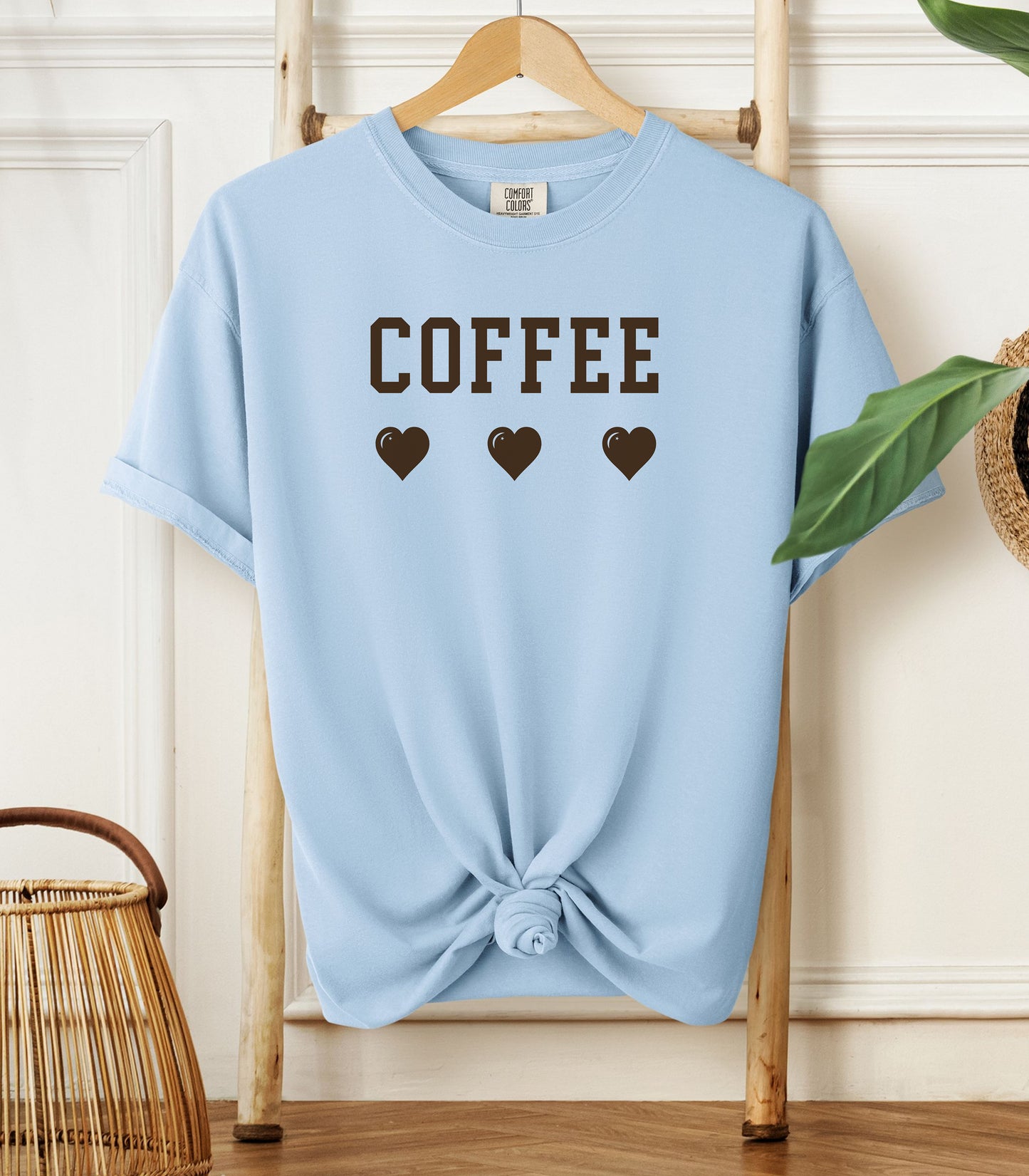 Chambray light blue Comfort Colors t-shirt featuring bold brown varsity-style “COFFEE” text above three matching heart icons. Simple and cute design that expresses love for coffee with a soft, cozy aesthetic—perfect for casual wear, coffee dates, or gifting to your favorite caffeine enthusiast.
