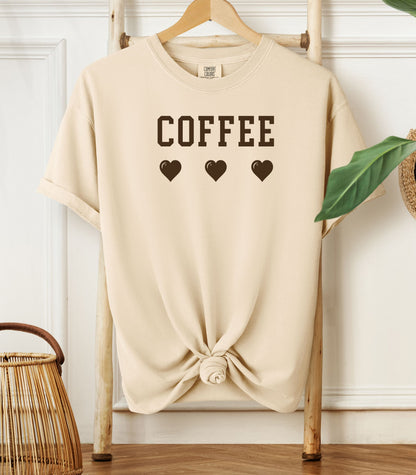Ivory Comfort Colors t-shirt featuring bold brown varsity-style “COFFEE” text above three matching heart icons. Simple and cute design that expresses love for coffee with a soft, cozy aesthetic—perfect for casual wear, coffee dates, or gifting to your favorite caffeine enthusiast.