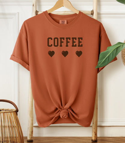 Yam Comfort Colors t-shirt featuring bold brown varsity-style “COFFEE” text above three matching heart icons. Simple and cute design that expresses love for coffee with a soft, cozy aesthetic—perfect for casual wear, coffee dates, or gifting to your favorite caffeine enthusiast.