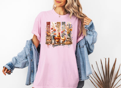 Country Life T-shirt in blossom pink featuring a vibrant farmhouse design with roosters, hens, eggs, sunflowers, butterflies, and rustic wooden crates. The artwork is accented with vertical watercolor panels and labeled elements like “Eggs,” “Butterflies,” “Chickens,” and “Sunflowers,” capturing the charm of rural living and barnyard beauty. Perfect for farm lovers and country style enthusiasts.