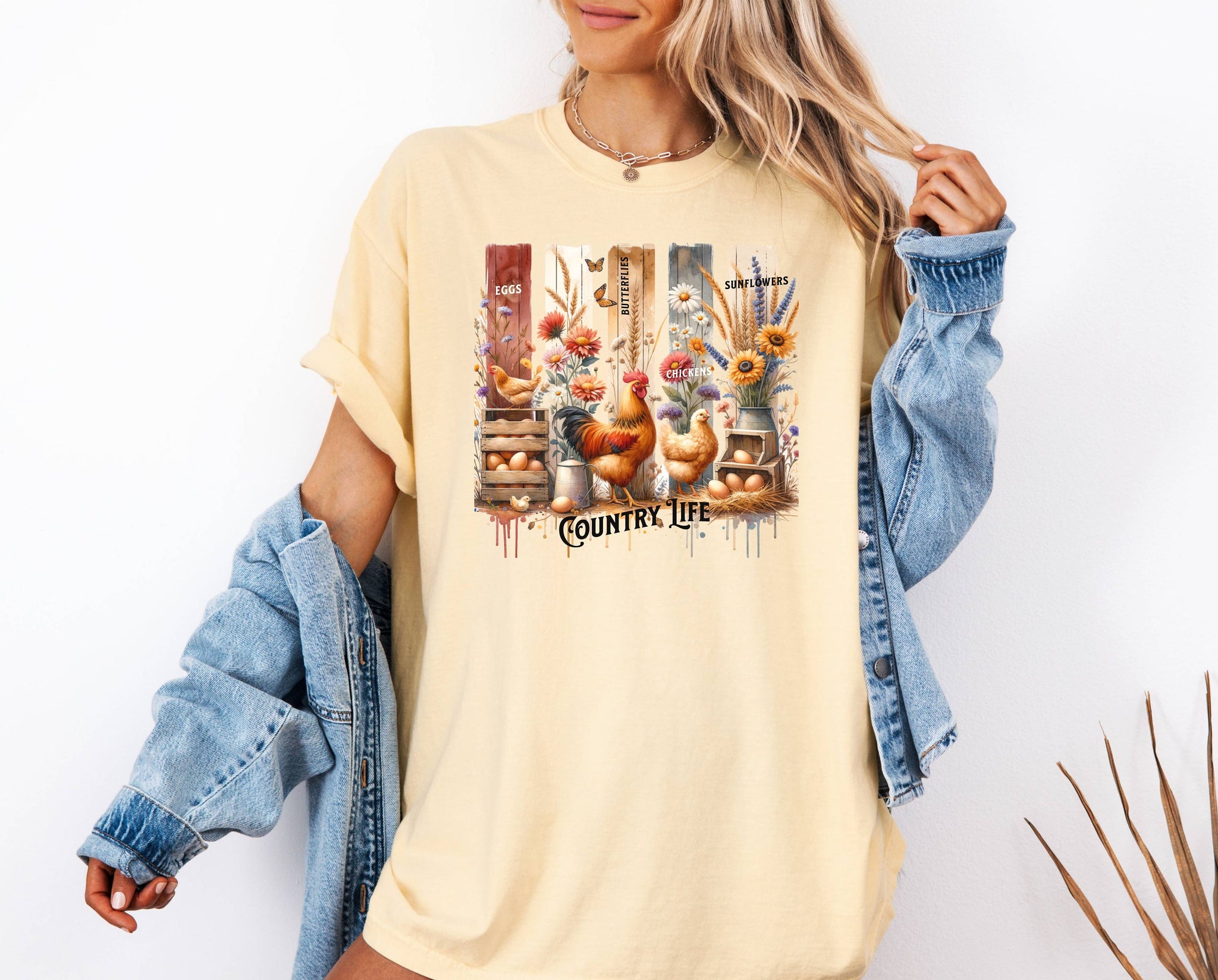Country Life T-shirt in soft butter featuring a vibrant farmhouse design with roosters, hens, eggs, sunflowers, butterflies, and rustic wooden crates. The artwork is accented with vertical watercolor panels and labeled elements like “Eggs,” “Butterflies,” “Chickens,” and “Sunflowers,” capturing the charm of rural living and barnyard beauty. Perfect for farm lovers and country style enthusiasts.