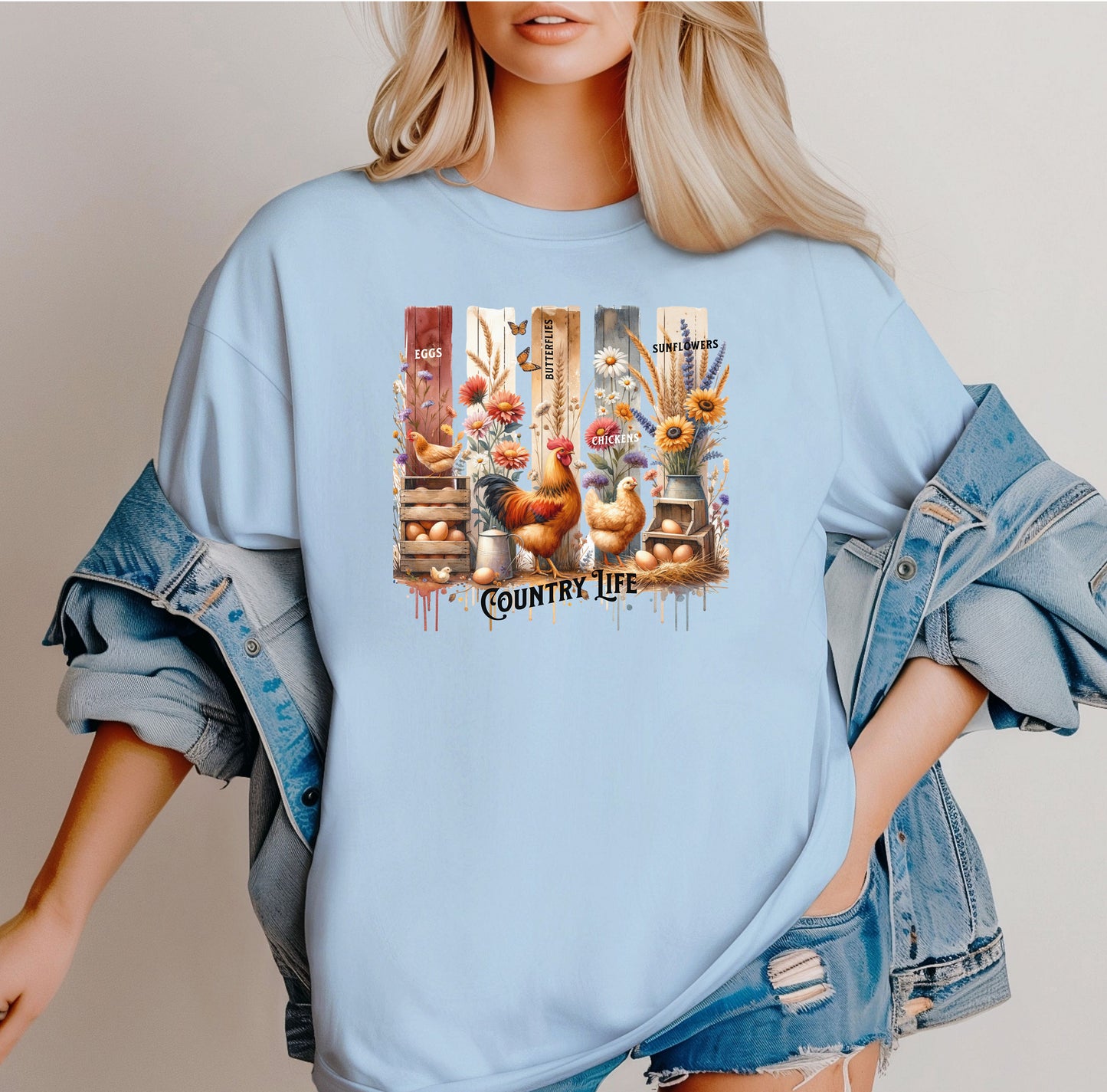 Country Life T-shirt in chambray light blue featuring a vibrant farmhouse design with roosters, hens, eggs, sunflowers, butterflies, and rustic wooden crates. The artwork is accented with vertical watercolor panels and labeled elements like “Eggs,” “Butterflies,” “Chickens,” and “Sunflowers,” capturing the charm of rural living and barnyard beauty. Perfect for farm lovers and country style enthusiasts.