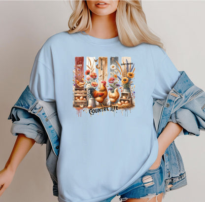 Country Life T-shirt in chambray light blue featuring a vibrant farmhouse design with roosters, hens, eggs, sunflowers, butterflies, and rustic wooden crates. The artwork is accented with vertical watercolor panels and labeled elements like “Eggs,” “Butterflies,” “Chickens,” and “Sunflowers,” capturing the charm of rural living and barnyard beauty. Perfect for farm lovers and country style enthusiasts.