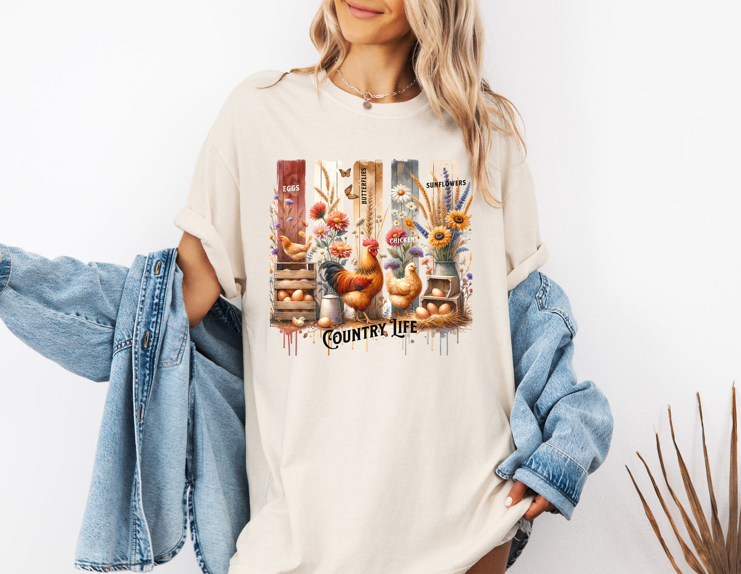 Country Life T-shirt in ivory featuring a vibrant farmhouse design with roosters, hens, eggs, sunflowers, butterflies, and rustic wooden crates. The artwork is accented with vertical watercolor panels and labeled elements like “Eggs,” “Butterflies,” “Chickens,” and “Sunflowers,” capturing the charm of rural living and barnyard beauty. Perfect for farm lovers and country style enthusiasts.