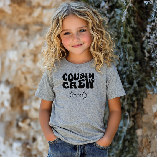 Smiling toddler wearing a Athletic Heather gray T-shirt featuring a bold black "Cousin Crew" design with “Your Name” in a cursive font underneath. The shirt has a casual fit and is styled with denim shorts, creating a playful and adorable look perfect for family events or matching cousin outfits.
