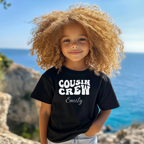 Smiling toddler wearing a Black T-shirt featuring a bold white "Cousin Crew" design with “Your Name” in a cursive font underneath. The shirt has a casual fit and is styled with denim shorts, creating a playful and adorable look perfect for family events or matching cousin outfits.