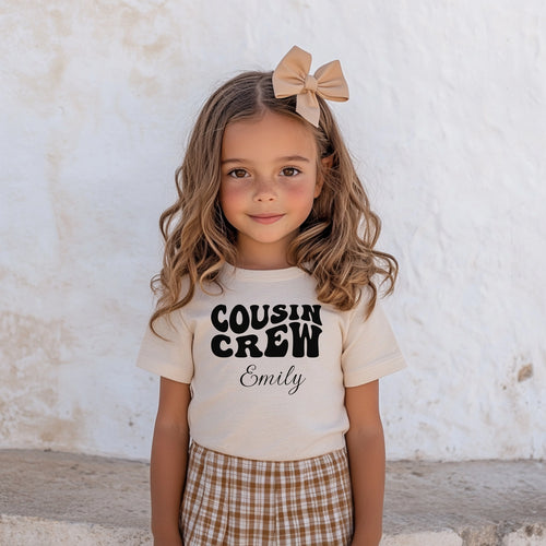 Smiling toddler wearing a Natural T-shirt featuring a bold black "Cousin Crew" design with “Your Name” in a cursive font underneath. The shirt has a casual fit and is styled with a cute plaid skirt, creating a playful and adorable look perfect for family events or matching cousin outfits.