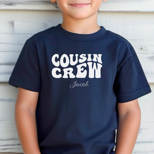 Smiling toddler wearing a Navy T-shirt featuring a bold white "Cousin Crew" design with “Your Name” in a cursive font underneath. The shirt has a casual fit and is styled with denim shorts, creating a playful and adorable look perfect for family events or matching cousin outfits.