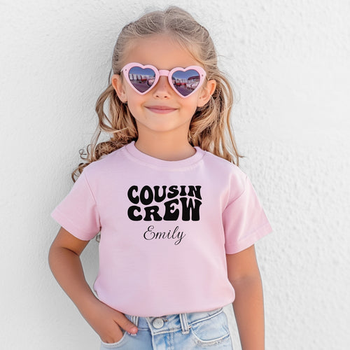 Smiling toddler wearing a Pink T-shirt featuring a bold black "Cousin Crew" design with “Your Name” in a cursive font underneath. The shirt has a casual fit and is styled with denim jeans, creating a playful and adorable look perfect for family events or matching cousin outfits.