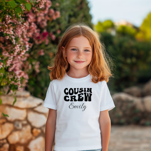 Smiling toddler wearing a White T-shirt featuring a bold black "Cousin Crew" design with “Your Name” in a cursive font underneath. The shirt has a casual fit and is styled with denim jeans, creating a playful and adorable look perfect for family events or matching cousin outfits.