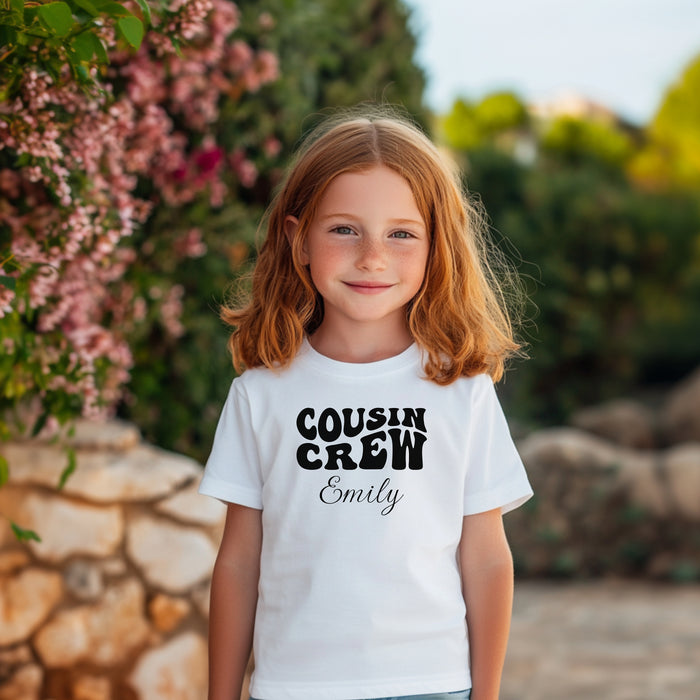 Smiling toddler wearing a White T-shirt featuring a bold black "Cousin Crew" design with “Your Name” in a cursive font underneath. The shirt has a casual fit and is styled with denim jeans, creating a playful and adorable look perfect for family events or matching cousin outfits.