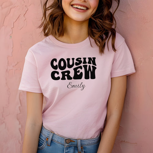 Smiling child wearing a pink youth T-shirt with bold black "Cousin Crew" text in a playful font and personalized with “Your Name” in elegant script below. The shirt is paired with jeans, creating a fun and youthful vibe perfect for family gatherings or matching cousin outfits.