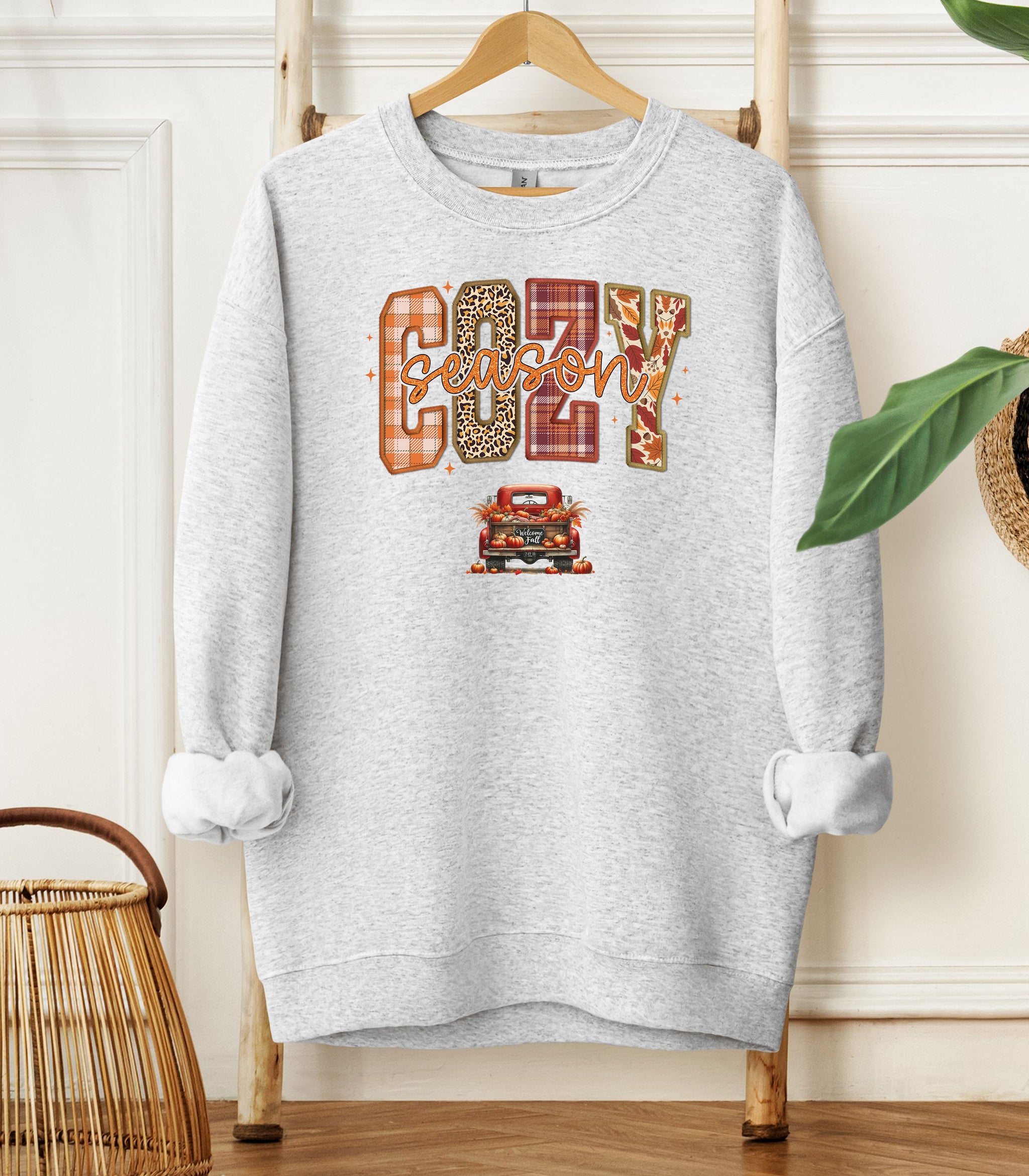 Ash Gildan sweatshirt featuring a colorful “Cozy Season” design with bold patterned letters—plaid, leopard, gingham, and fall leaves—and a retro red truck filled with pumpkins and sunflowers below. A festive and warm autumn look perfect for embracing pumpkin spice vibes, crisp air, and cozy fall days.
