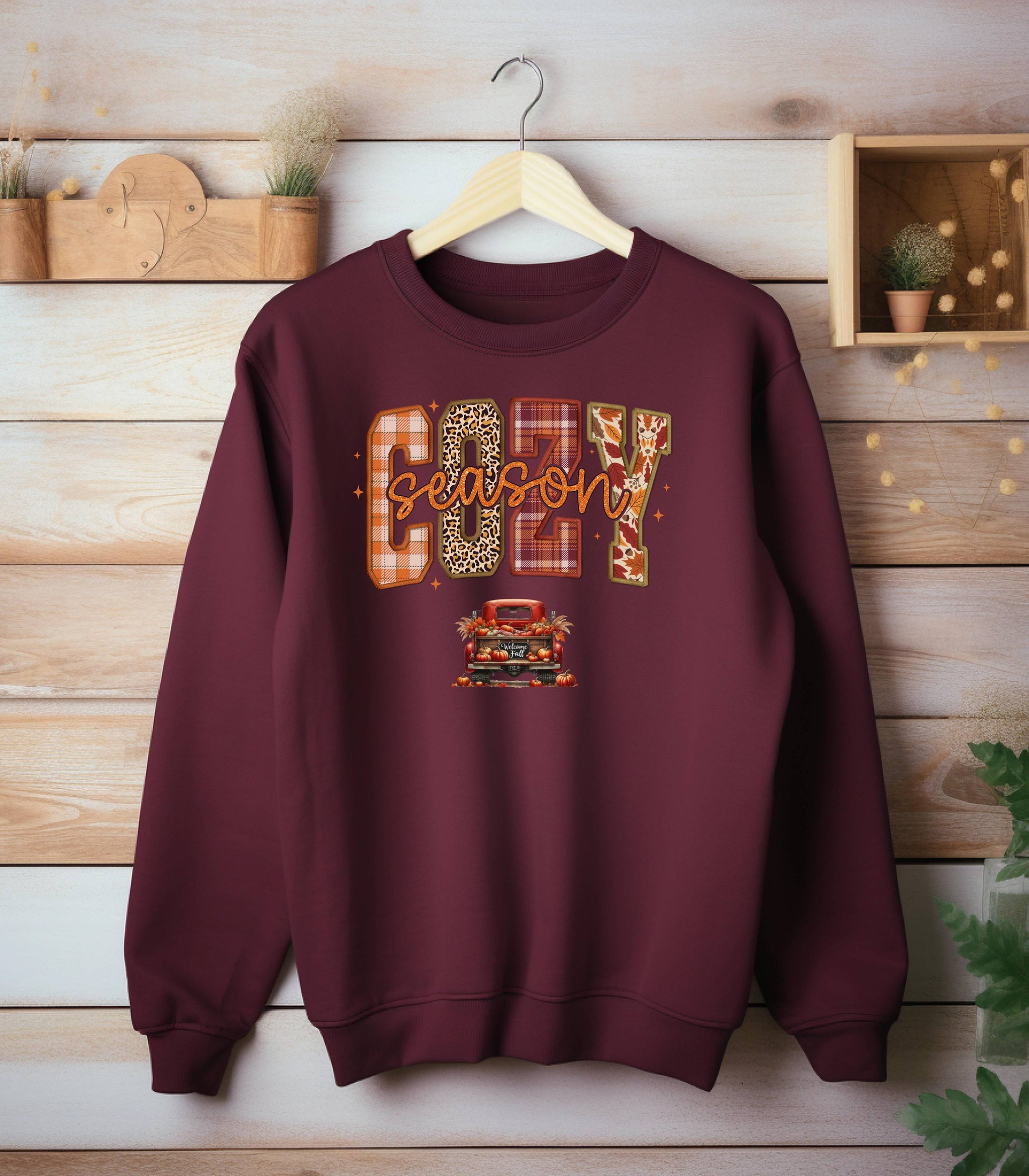 Maroon Gildan sweatshirt featuring a colorful “Cozy Season” design with bold patterned letters—plaid, leopard, gingham, and fall leaves—and a retro red truck filled with pumpkins and sunflowers below. A festive and warm autumn look perfect for embracing pumpkin spice vibes, crisp air, and cozy fall days.