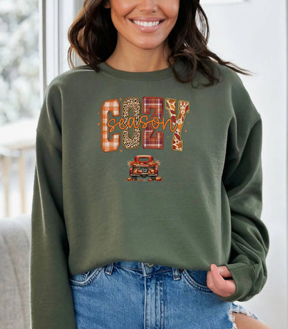 Military Green Gildan sweatshirt featuring a colorful “Cozy Season” design with bold patterned letters—plaid, leopard, gingham, and fall leaves—and a retro red truck filled with pumpkins and sunflowers below. A festive and warm autumn look perfect for embracing pumpkin spice vibes, crisp air, and cozy fall days.