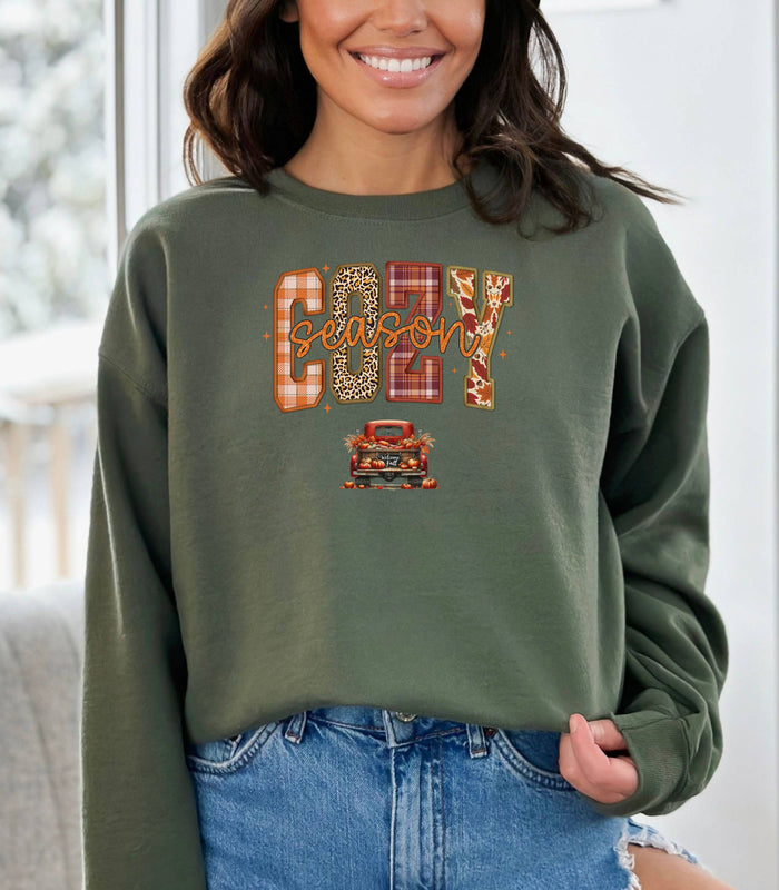 Military Green Gildan sweatshirt featuring a colorful “Cozy Season” design with bold patterned letters—plaid, leopard, gingham, and fall leaves—and a retro red truck filled with pumpkins and sunflowers below. A festive and warm autumn look perfect for embracing pumpkin spice vibes, crisp air, and cozy fall days.
