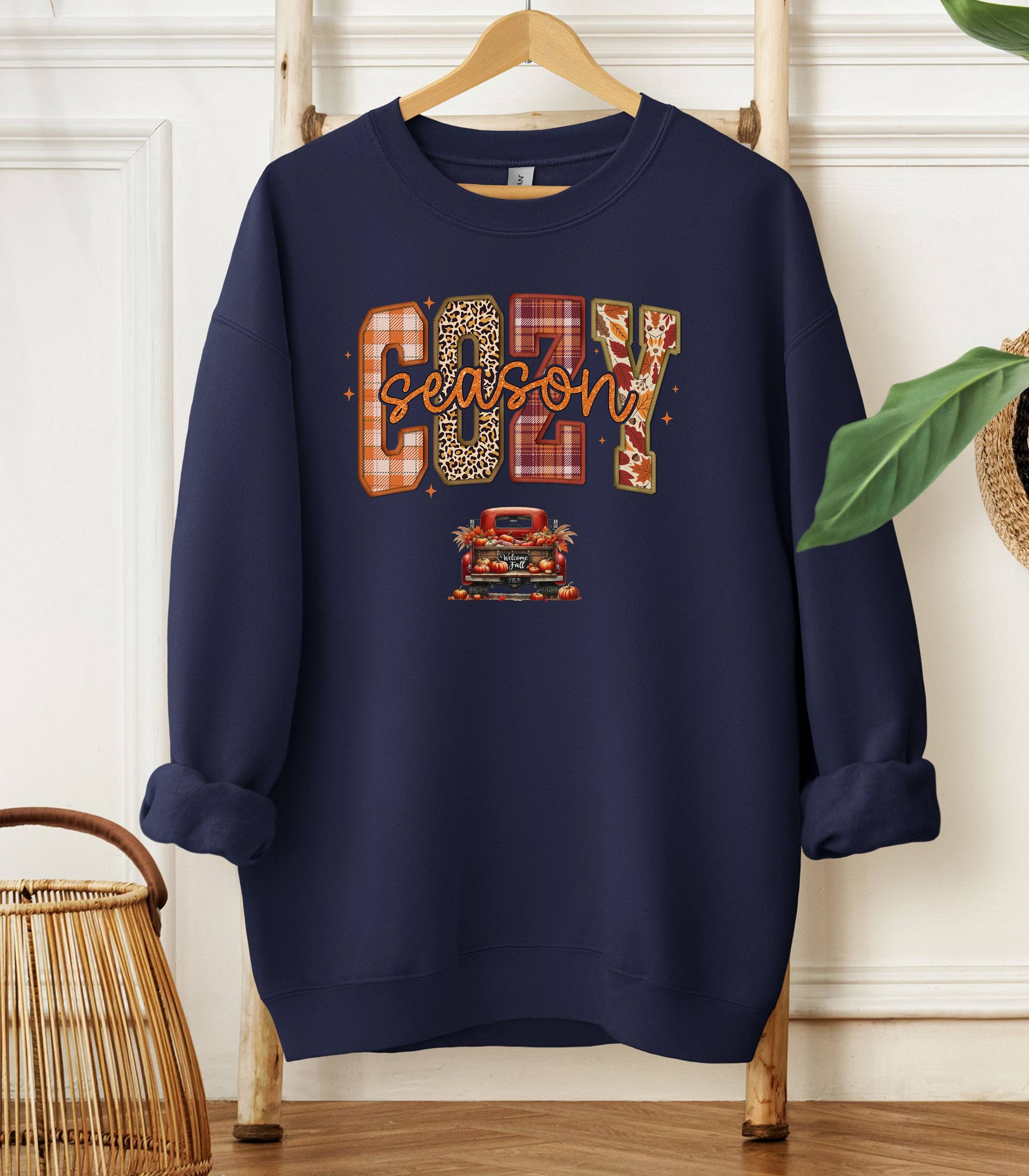 Navy Gildan sweatshirt featuring a colorful “Cozy Season” design with bold patterned letters—plaid, leopard, gingham, and fall leaves—and a retro red truck filled with pumpkins and sunflowers below. A festive and warm autumn look perfect for embracing pumpkin spice vibes, crisp air, and cozy fall days.