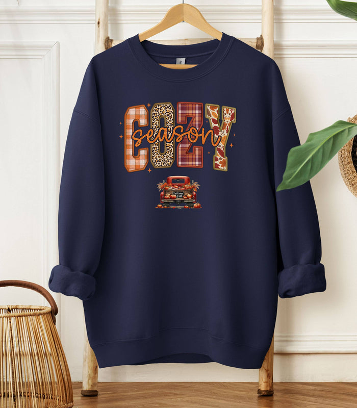 Navy Gildan sweatshirt featuring a colorful “Cozy Season” design with bold patterned letters—plaid, leopard, gingham, and fall leaves—and a retro red truck filled with pumpkins and sunflowers below. A festive and warm autumn look perfect for embracing pumpkin spice vibes, crisp air, and cozy fall days.
