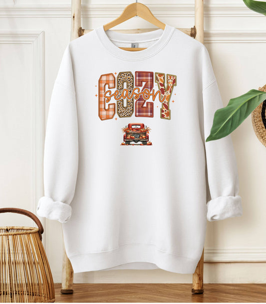 White Gildan sweatshirt featuring a colorful “Cozy Season” design with bold patterned letters—plaid, leopard, gingham, and fall leaves—and a retro red truck filled with pumpkins and sunflowers below. A festive and warm autumn look perfect for embracing pumpkin spice vibes, crisp air, and cozy fall days.