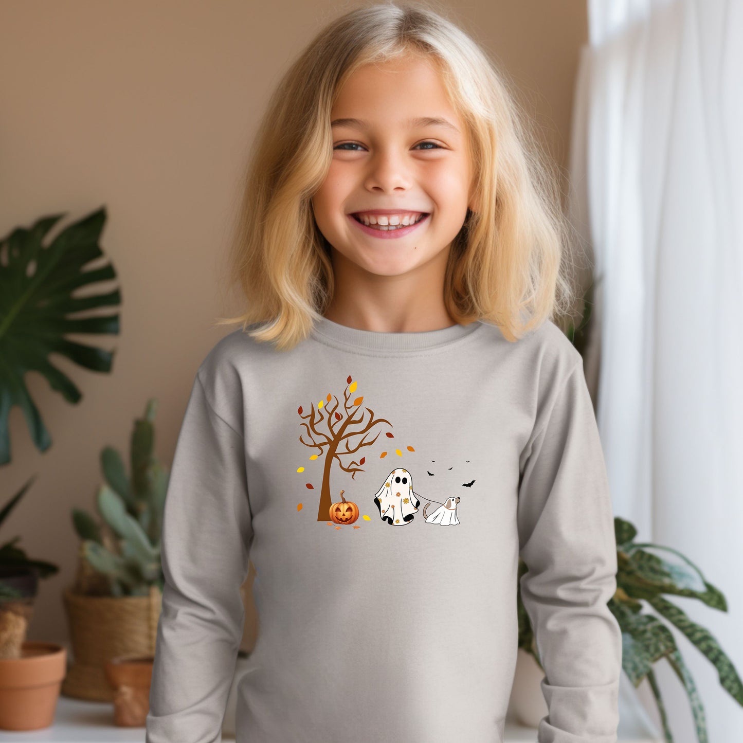 Adorable toddler wearing a Heather long-sleeve T-shirt with a cute Halloween design featuring a spooky tree with falling leaves, a smiling jack-o'-lantern, and two ghost figures—one child ghost and one ghost dog. A playful and heartwarming fall design perfect for Halloween festivities and toddler apparel.