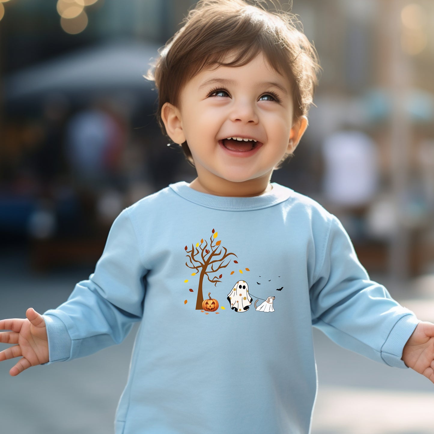 Adorable toddler wearing a light blue long-sleeve T-shirt with a cute Halloween design featuring a spooky tree with falling leaves, a smiling jack-o'-lantern, and two ghost figures—one child ghost and one ghost dog. A playful and heartwarming fall design perfect for Halloween festivities and toddler apparel.