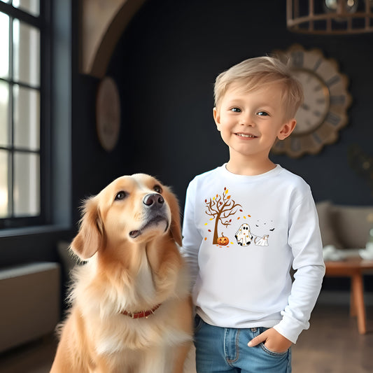 Adorable toddler wearing a white long-sleeve T-shirt with a cute Halloween design featuring a spooky tree with falling leaves, a smiling jack-o'-lantern, and two ghost figures—one child ghost and one ghost dog. A playful and heartwarming fall design perfect for Halloween festivities and toddler apparel.